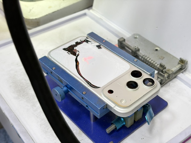 工程師將 iPhone 17 Pro Max 固定在藍色夾具上，使用熱風槍加熱並用特製刀片剔除機背碎玻璃